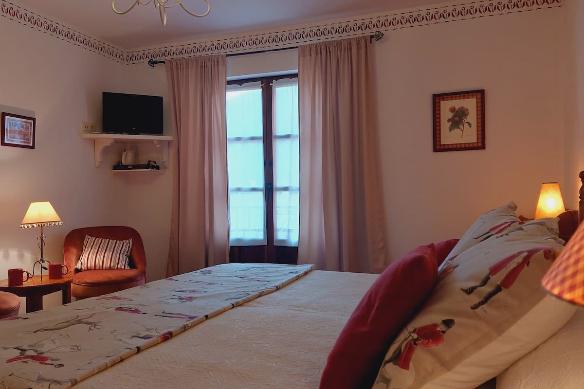 Una habitación de hotel con cama, una silla, televisor, ventana con cortinas y lámpara.