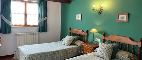A room with two beds, wooden headboards, a window with curtains, a wall-mounted lamp, and framed pictures.