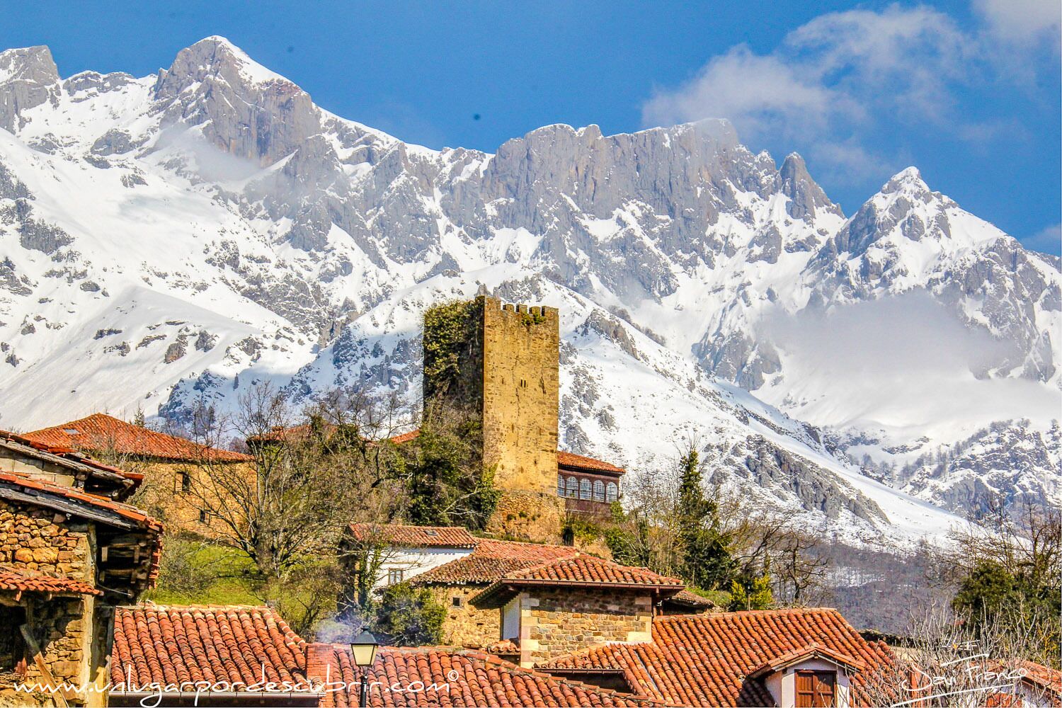 A historic stone tower amidst snow-covered mountains, with traditional red-tiled buildings in the foreground.