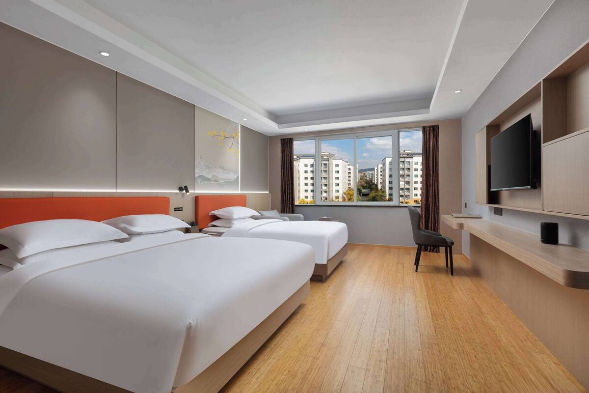 A hotel room with two beds, a TV, a desk, and a large window with a city view.