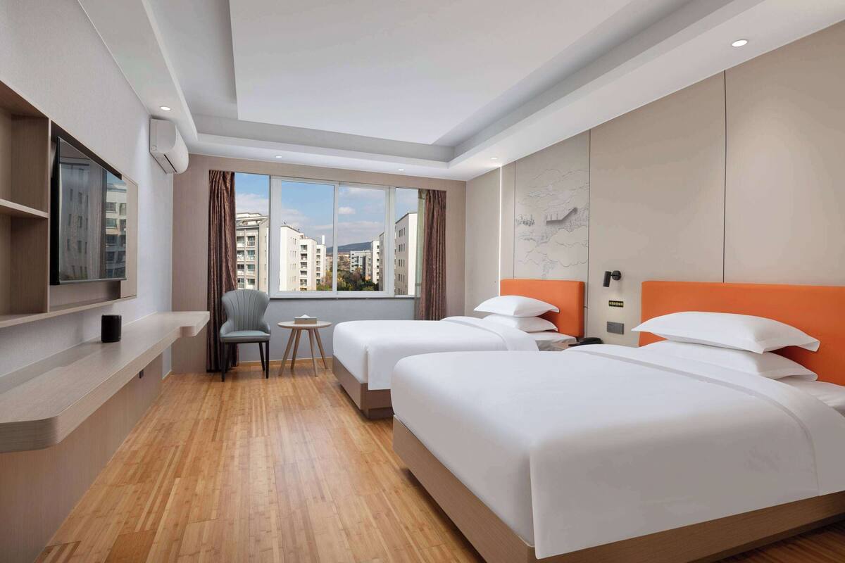 A hotel room with two beds, a TV, a small table, and a window with a city view.