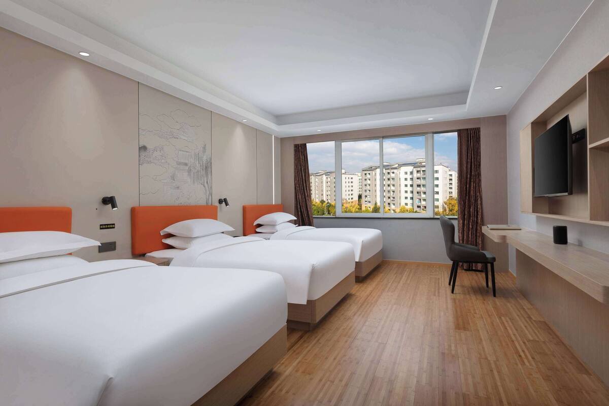 A hotel room with three beds, a desk, a chair, and a large window with a city view.