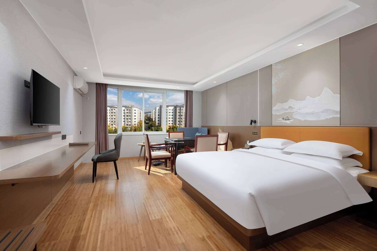 A modern hotel room with a large bed, a desk with a chair, a flat-screen TV, and a large window offering a city view.