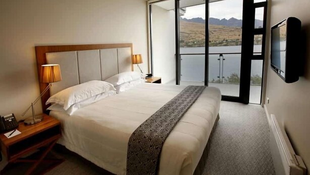 Una habitación de hotel con una cama grande, una mesita de noche, una lámpara, un televisor y un balcón con vistas a montañas y un lago.
