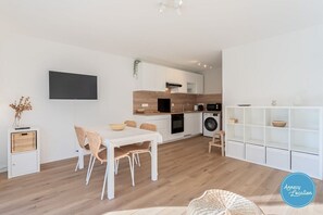 A modern kitchen with white cabinetry, a flat-screen TV, and a dining area with a white table and wicker chairs. - Lakefront T2 Apartment – Rive du Lac (Menthon-Saint-Bernard)