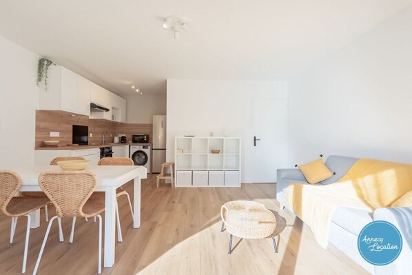 A modern, well-lit apartment with a dining area, kitchenette, and a living space with a sofa and cushions. - Lakefront T2 Apartment – Rive du Lac (Menthon-Saint-Bernard)