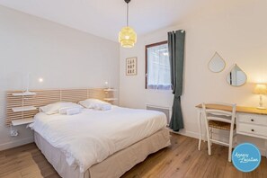 Un dormitorio con cama, mesita de noche, silla, escritorio, lámpara, ventana con cortinas y un espejo montado en la pared.