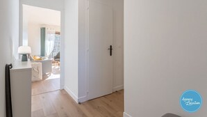 A hallway leading to a room with a door, a lamp, and a view of a balcony with a chair and a table.