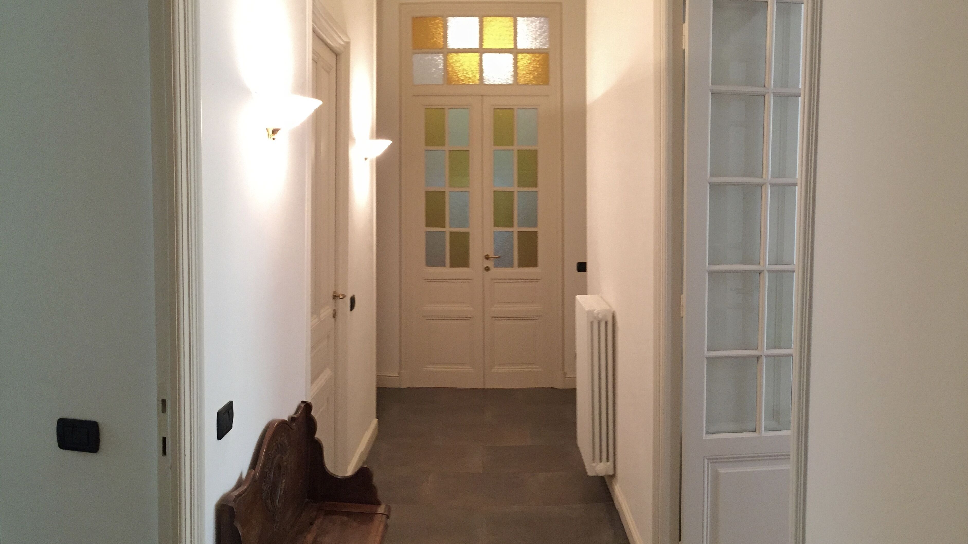 A narrow hallway with a double door at the end, a radiator on the right, and a bench on the left.