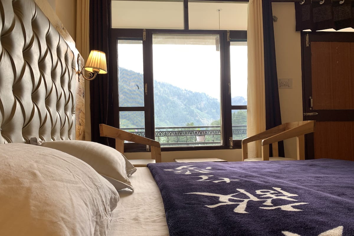 A bed with a headboard, a window with a view of mountains, and a lamp on the wall.
