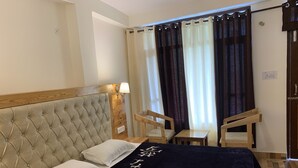 A hotel room with a tufted headboard, a bed with white linens, a dark throw blanket with white text, a wooden nightstand, a wall-mounted lamp, a small wooden table with a chair, and a window with curtains. - Jungle by sturmfrei Manali (Manali)