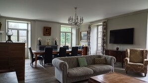 Un salon spacieux avec un canapé, une table basse et des fauteuils. On y trouve également un coin repas avec une table et des chaises, ainsi qu’un lustre suspendu au plafond. Un téléviseur est fixé au mur.