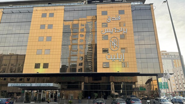 Un bâtiment moderne doté d’une grande façade en verre réfléchissant, d’une signalétique en arabe et d’une entrée bien visible, avec une voiture garée à l’extérieur.