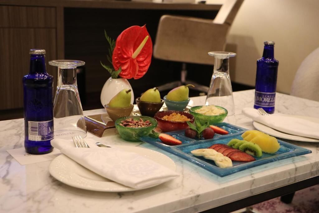 A breakfast table with various fruits, a vase with a red flower, and two bottles of water.