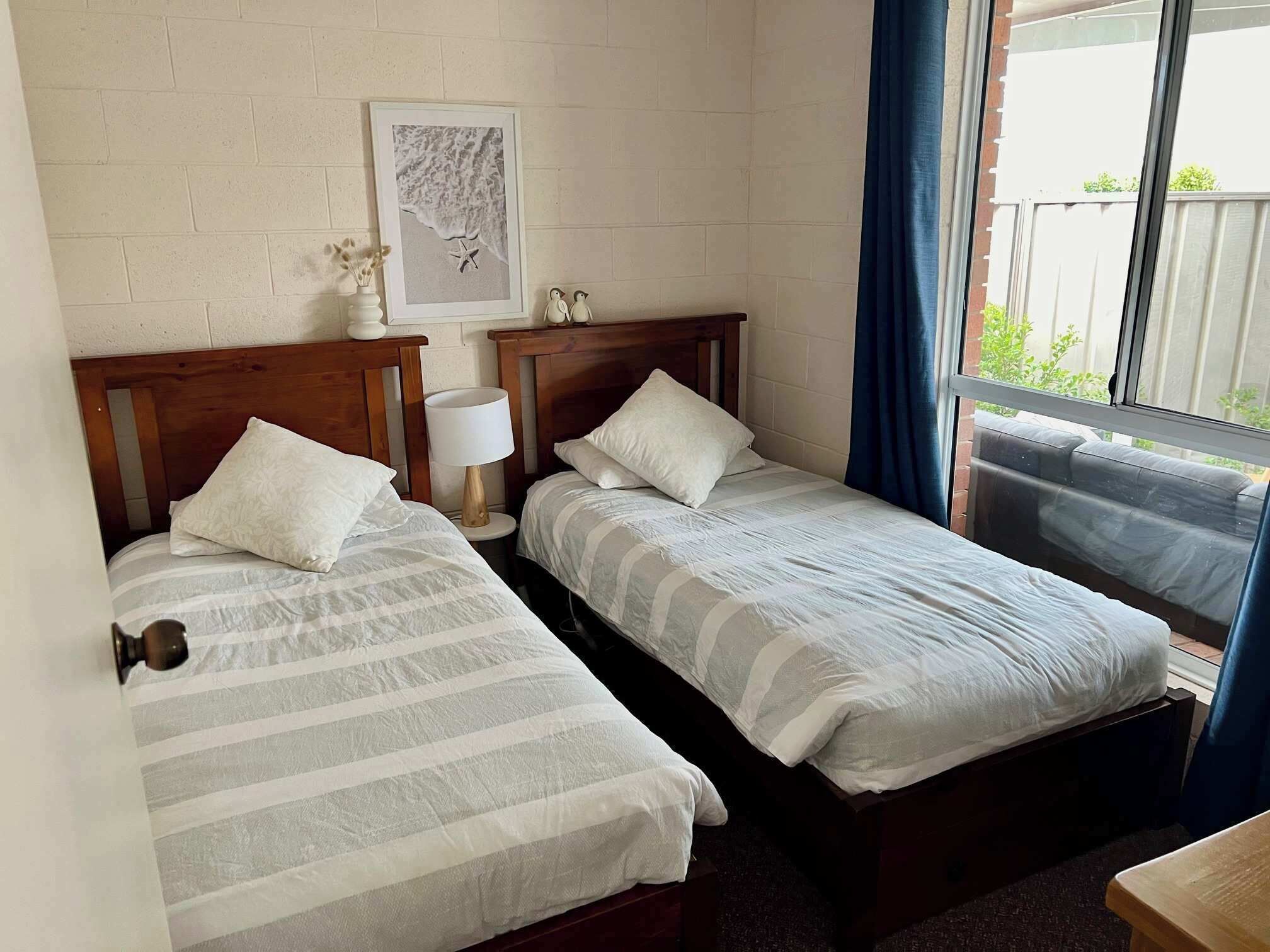 Une chambre avec deux lits simples, une petite table, une lampe, un tableau encadré au mur et une fenêtre avec des rideaux.