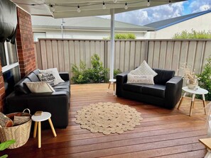 A covered outdoor seating area with two black sofas, a woven rug, and a small table with a bowl and flowers. - The J's - Pet Friendly Outside (Robe)