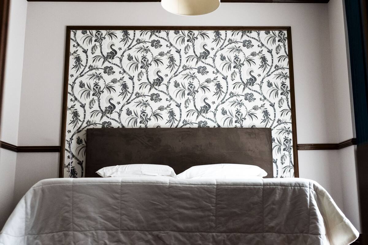 Ein Bett mit dunklem Kopfteil, weißer Bettwäsche und floralem Wandpapier an der Wand.