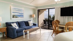 Une pièce avec un canapé bleu, un banc en osier, une table à manger avec des chaises, un téléviseur et une grande fenêtre donnant sur un beau paysage.