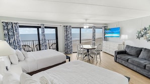 A hotel room with two beds, a dining area, a sofa, a TV, and a balcony with a view of the beach. - Vacation Time Oceanfront Deluxe Queen Room 4th Flr (Pawleys Island)