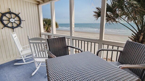 A covered outdoor seating area with wicker furniture, a white rocking chair, and a view of the beach. - Oceanfront Views King Room with Amenities (Pawleys Island)