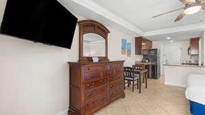 A room with a wooden dresser, a flat-screen TV, a ceiling fan, a dining area with chairs, and a kitchen area. - Dunes King Room Oceanfront Views (Pawleys Island)