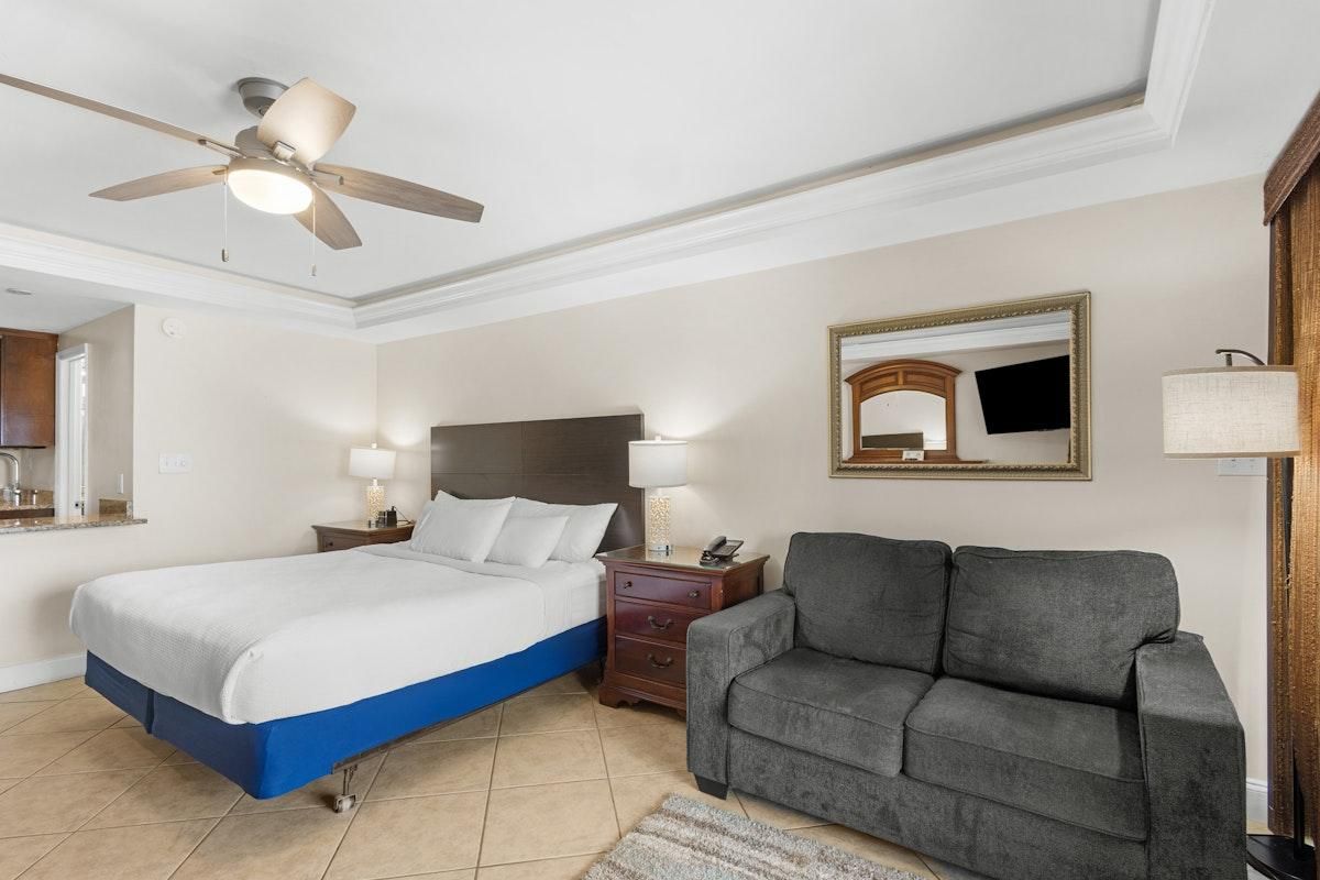 Hotellihuone, jossa on sänky, harmaa sohva, kattotuuletin, yöpöytä ja peili.