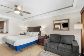 Hotellihuone, jossa on sänky, harmaa sohva, kattotuuletin, yöpöytä ja peili.