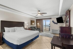 A hotel room with a large bed, a ceiling fan, a flat-screen TV, a dining table, and a view of the outdoors. - Dunes King Room Oceanfront Views (Pawleys Island)