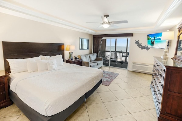Un dormitorio amplio con una cama grande, ventilador de techo, balcón con vista al mar y escritorio con lámpara.