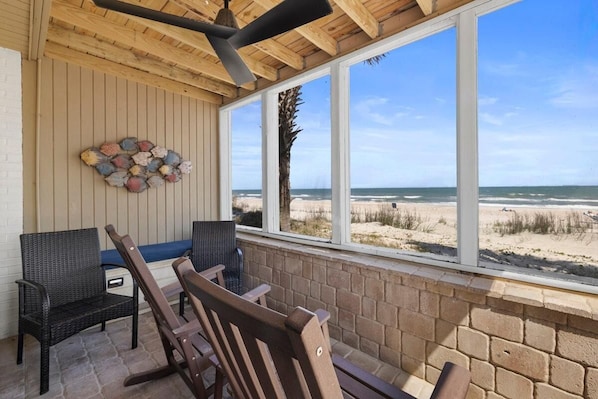 A covered outdoor seating area with wicker chairs and a table, overlooking a beach.