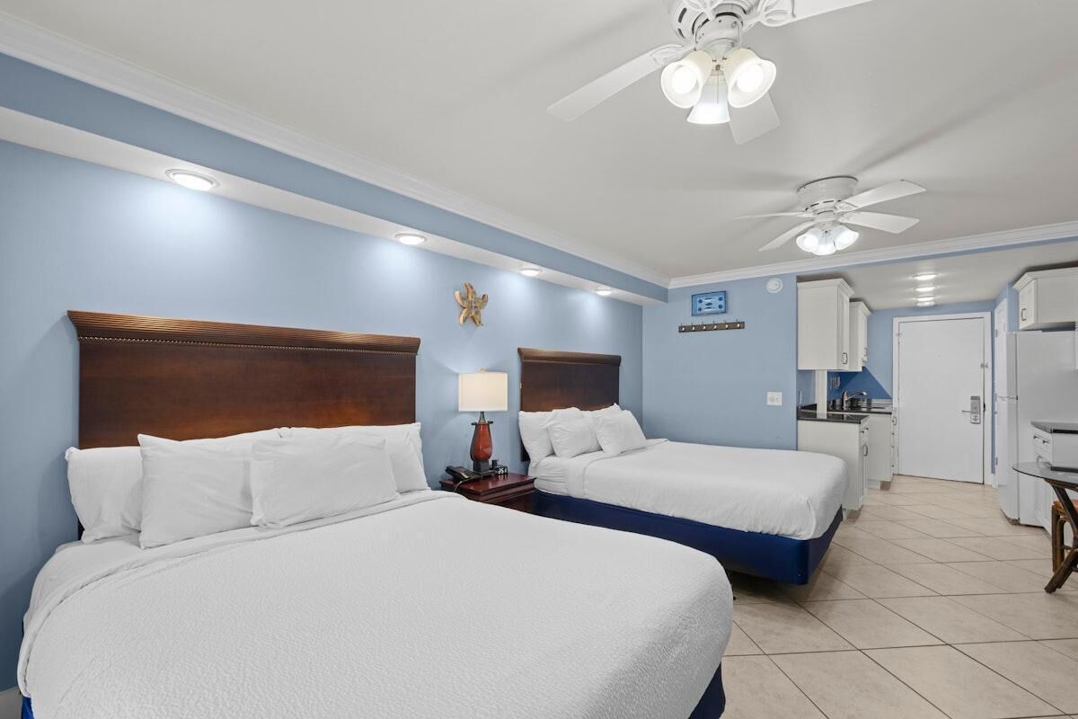 Een hotelkamer met twee bedden, een plafondventilator, een klein bureau met lamp en een kitchenette.