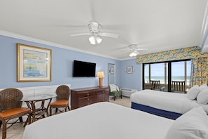 A hotel room with two beds, a television, a dining area with wicker chairs, a dresser, and a balcony with a view.