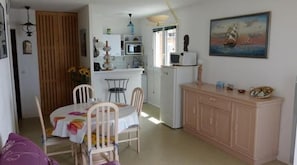 Een kleine keuken met een magnetron, spoelbak en kastjes. Er is een eethoek met een tafel en stoelen. Een schilderij hangt aan de muur.