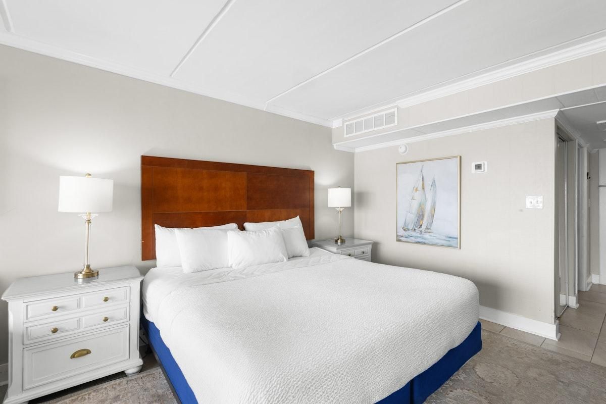 A hotel room with a large bed, two bedside tables, a nightstand, a painting on the wall, and a door leading to another room.
