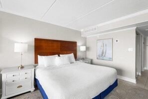 Una habitación de hotel con una cama grande, dos mesitas de noche, un buró, un cuadro en la pared y una puerta que da a otra habitación.