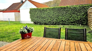Een houten tafel met stoelen, een rode bloempot en een haag in een tuin.