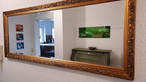 Een spiegel met een sierlijk gouden frame, reflecterend een kamer met een abstract schilderij in groen, een kast en een deur die leidt naar een andere kamer.