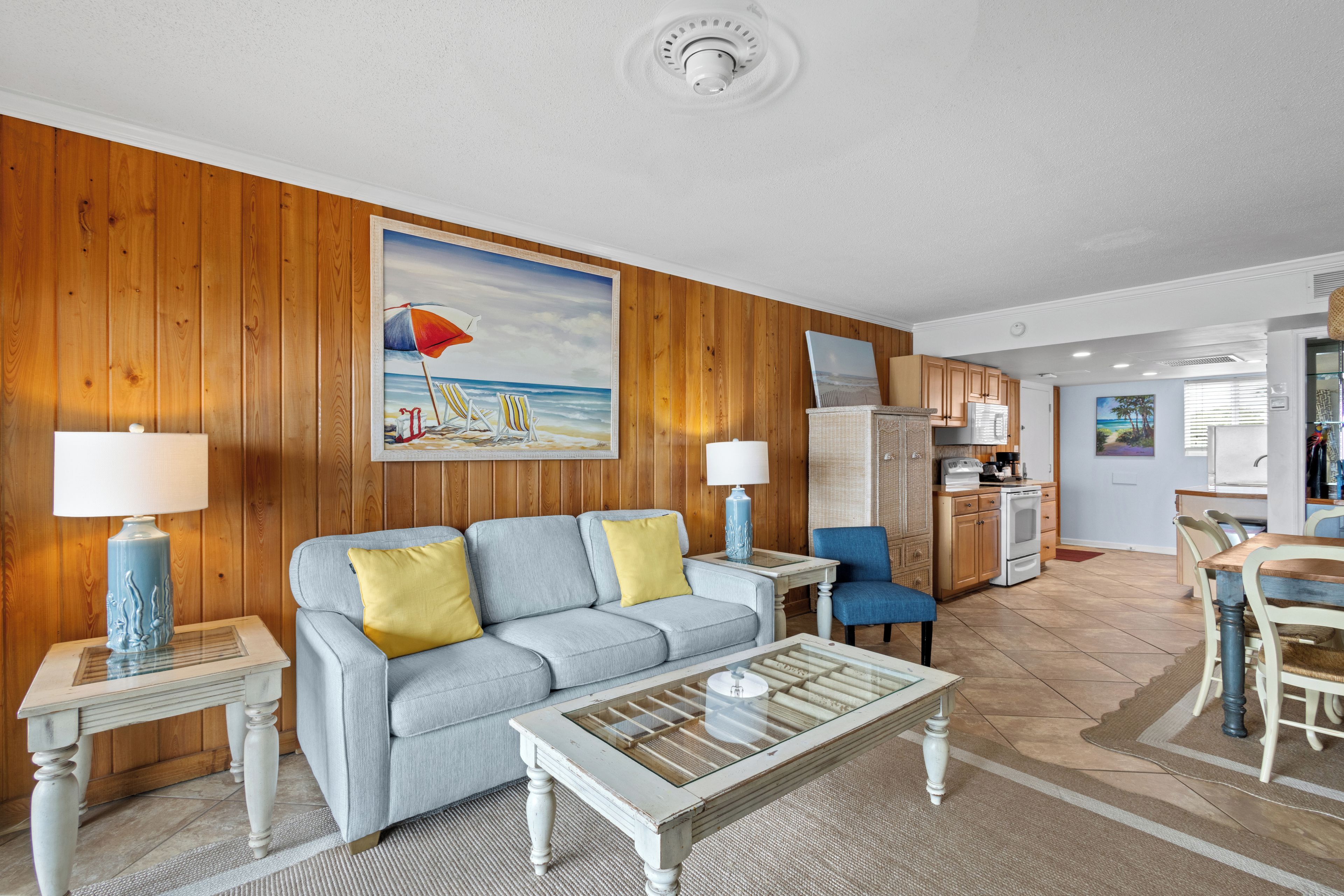 A living room with a sofa, coffee table, and lamps. There is a painting of a beach scene on the wall.