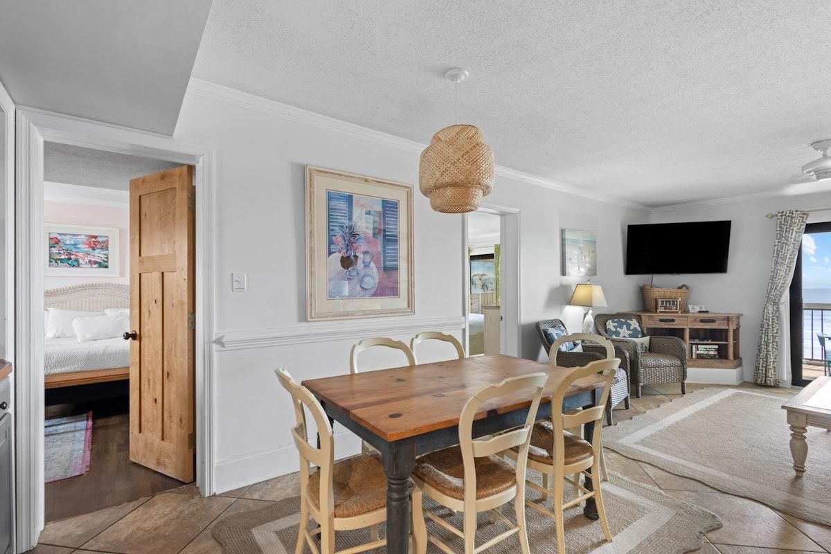A dining area with a wooden table and chairs, a hanging light fixture, and a television in the background.