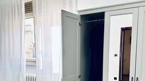 Ein Zimmer mit einem weißen Kleiderschrank, einem Heizkörper und einem Fenster mit durchsichtigen Vorhängen.