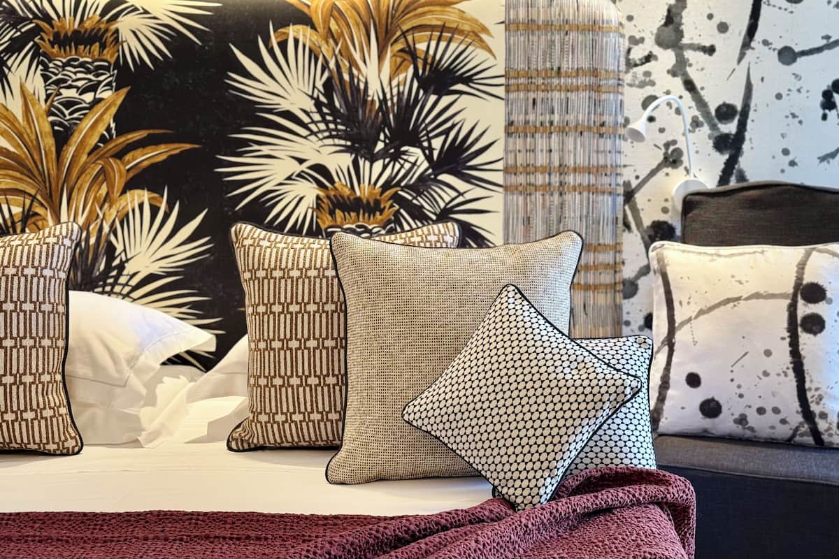 Una cama con varias almohadas, una colcha y una pared con una planta y un diseño abstracto.