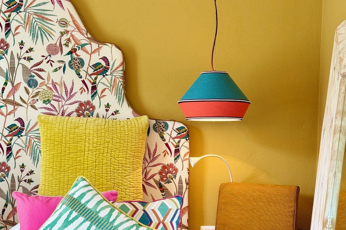 Un dormitorio acogedor con un cabecero floral, varios cojines de colores, una manta tejida, una mesita de noche de madera con una lámpara y una silla con una figura decorativa.