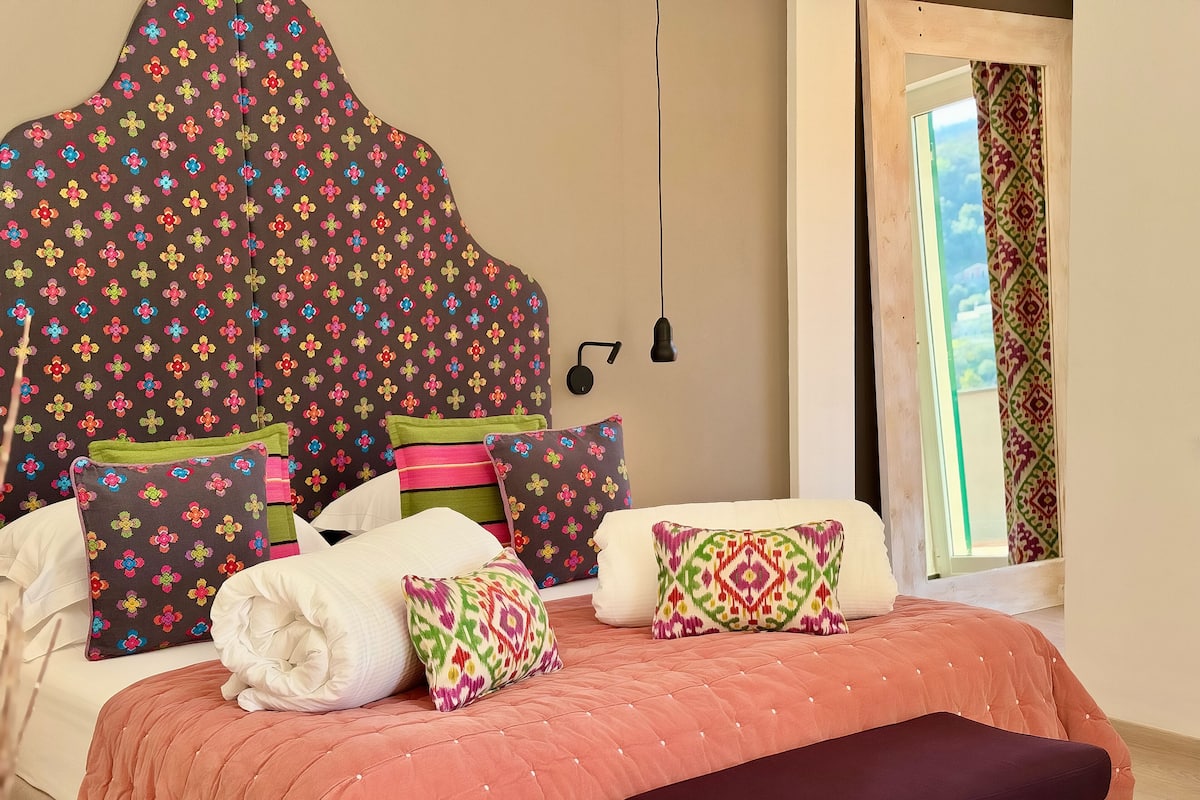 Un dormitorio con una cama grande, varios almohadones, un cabecero y una ventana con cortinas.