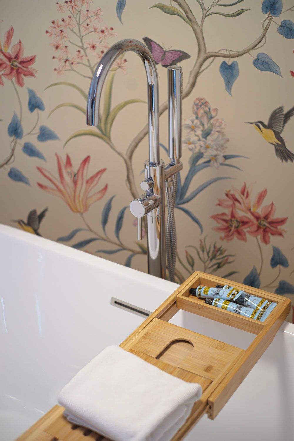 Ein Badezimmer mit einer weißen Badewanne, einem Holztablett mit einem Handtuch und floralem Wandpapier.