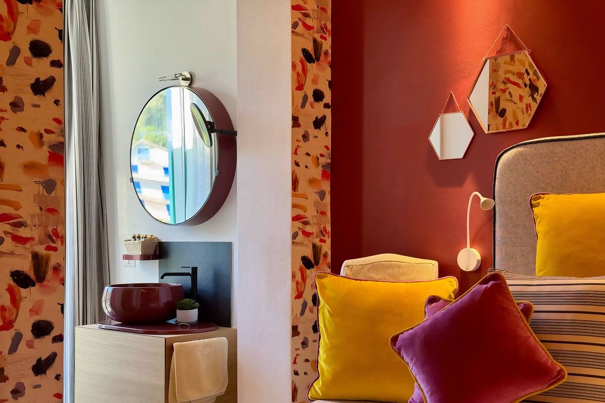 Una habitación con un espejo redondo, una mesita de noche con un cuenco rojo y una cama con almohadas amarillas y rojas.