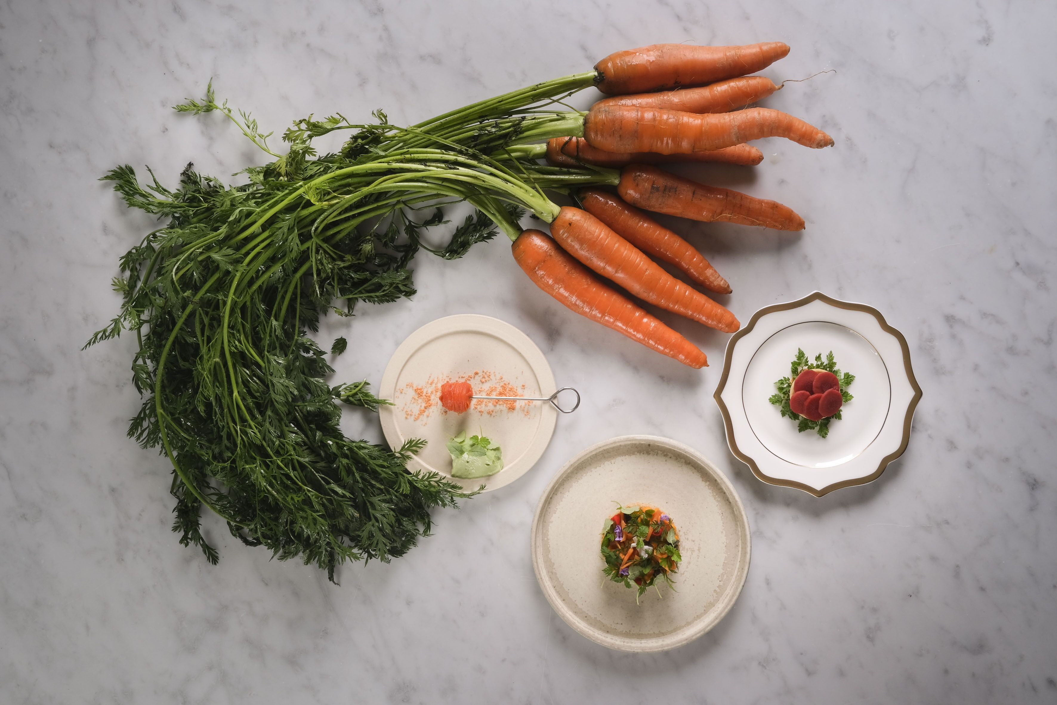 A top-down view of a kitchen counter with fresh carrots, parsley, and two plates with food.