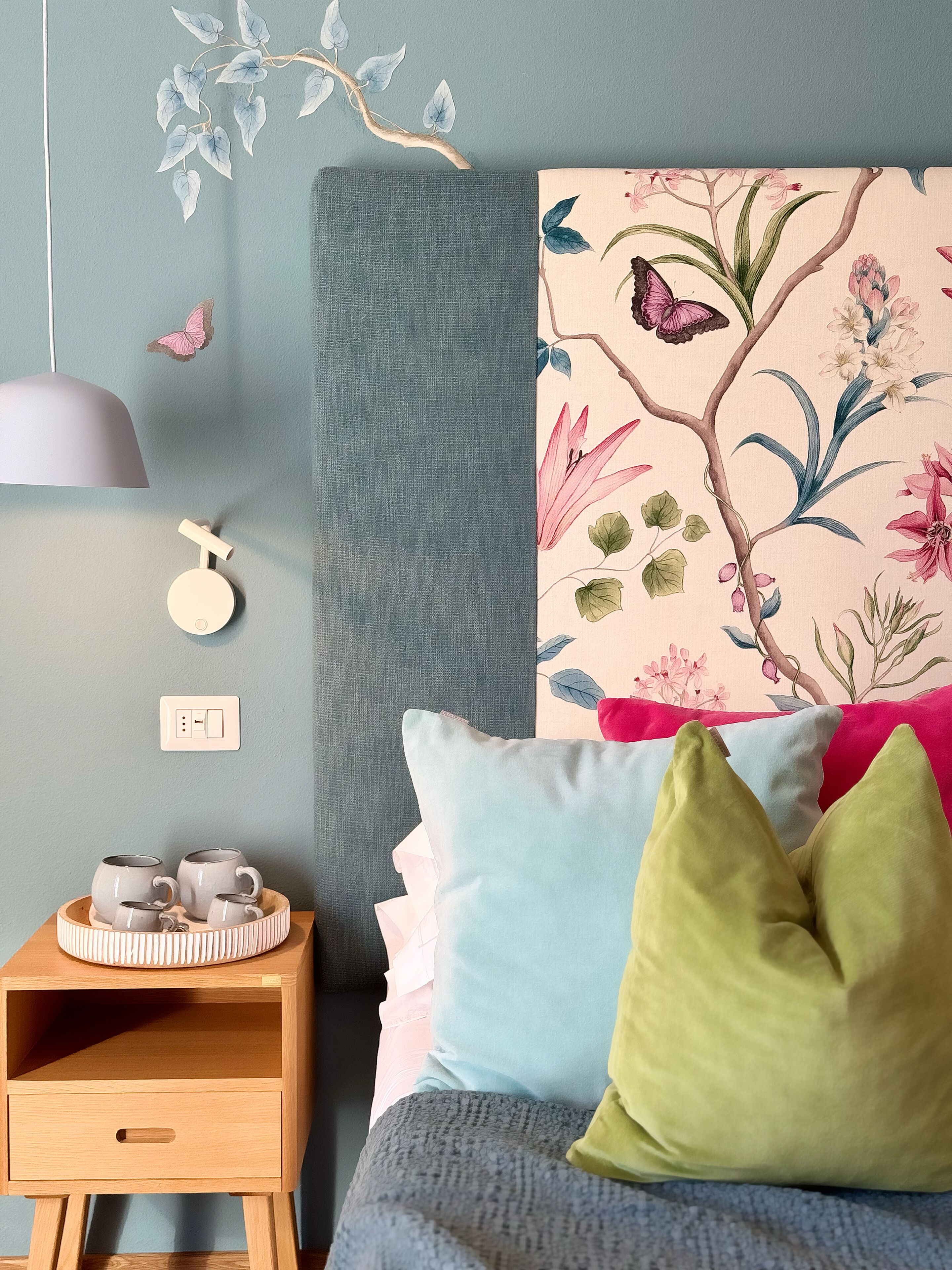 Et soveværelse med en seng, puder, et natbord, en vægmonteret lampe og et blomstermotiv.