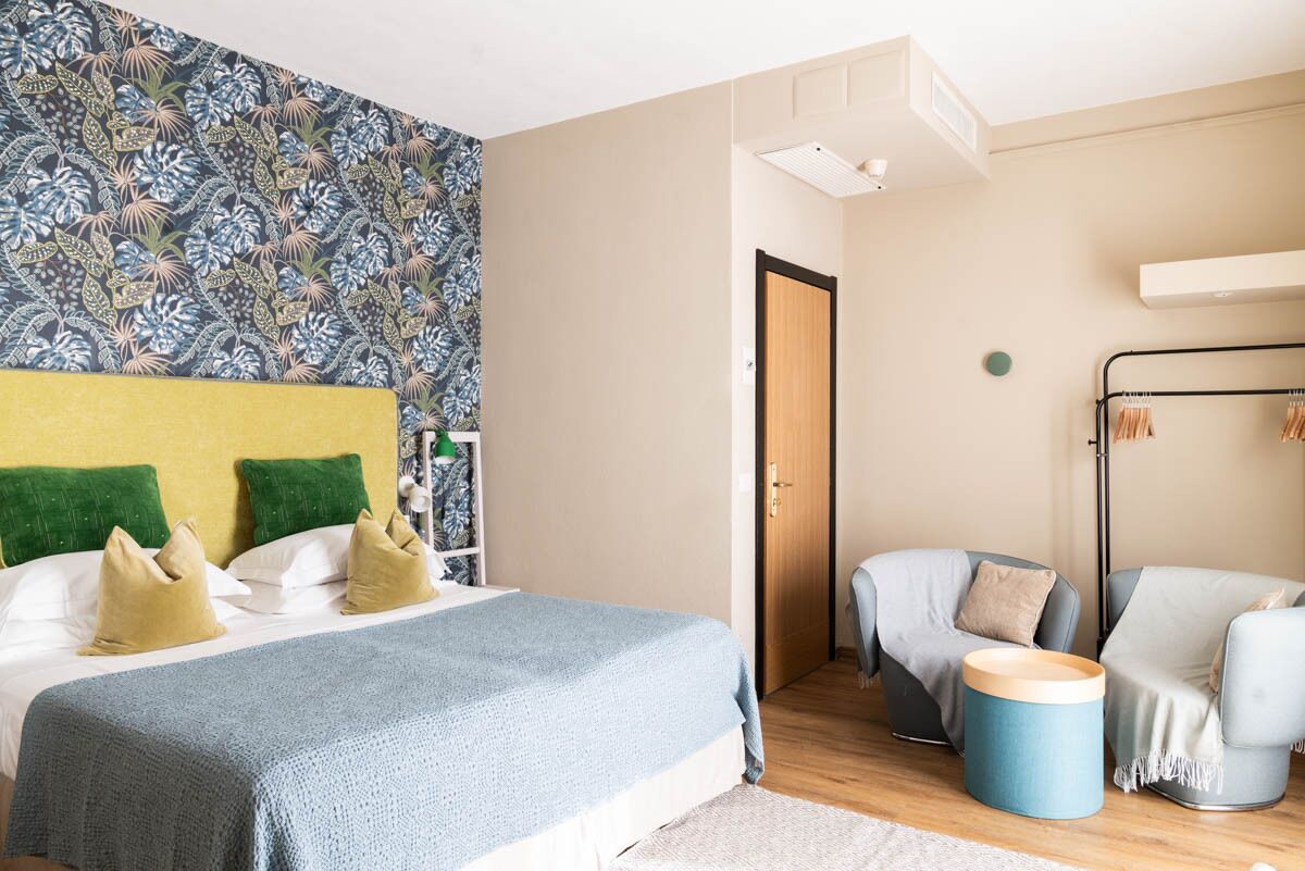 A hotel room with a bed, a wardrobe, a seating area with chairs, and a patterned wall.
