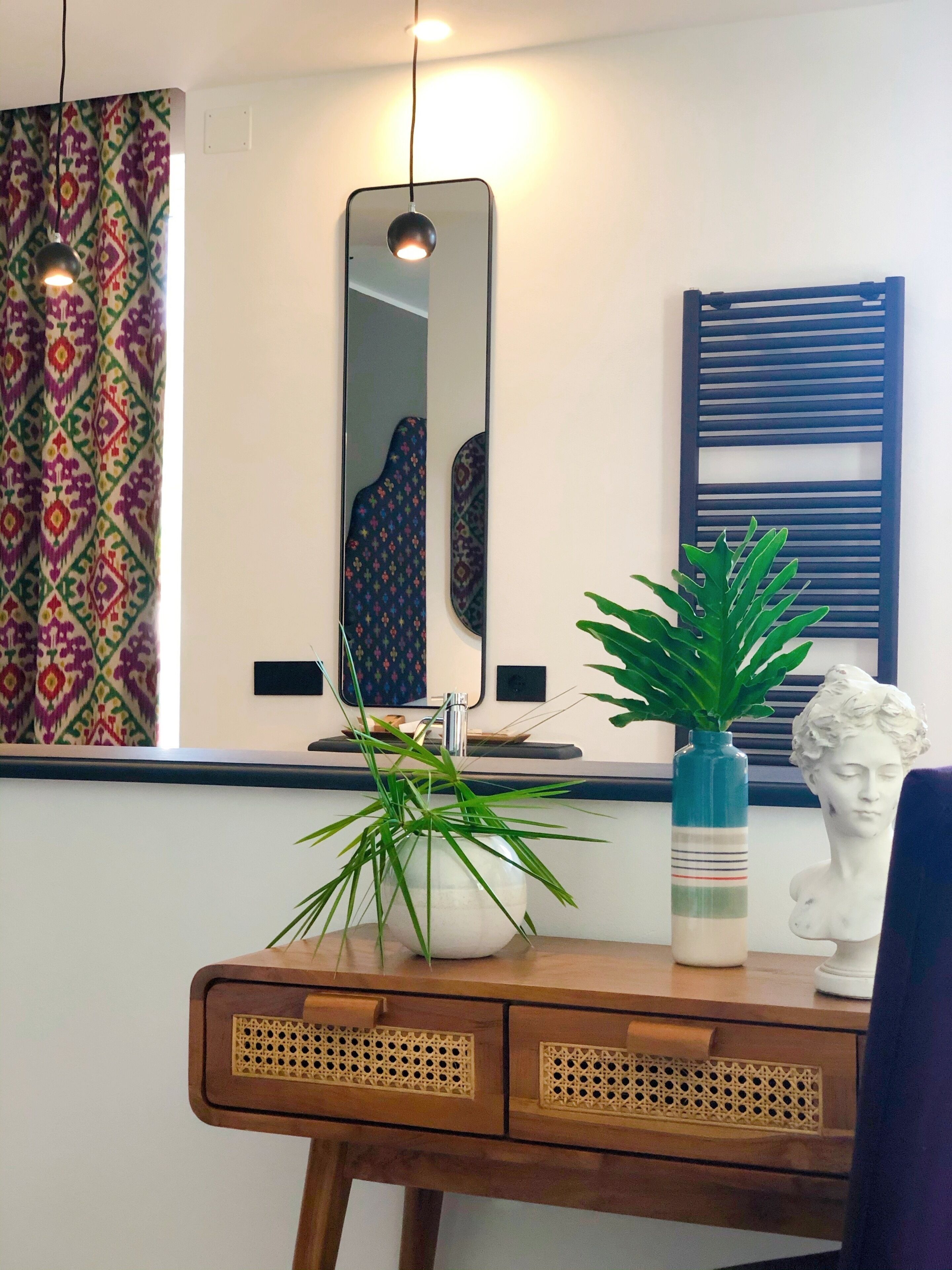 Une table en bois sur laquelle est posé un vase rempli de plantes vertes et une sculpture blanche. Un miroir est accroché au mur, au-dessus de la table.