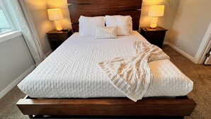 Una cama bien hecha con un edredón blanco acolchado, dos almohadas y una manta. Se ven dos lámparas de noche sobre mesitas de madera. También hay una decoración geométrica en la pared y una planta colgante.